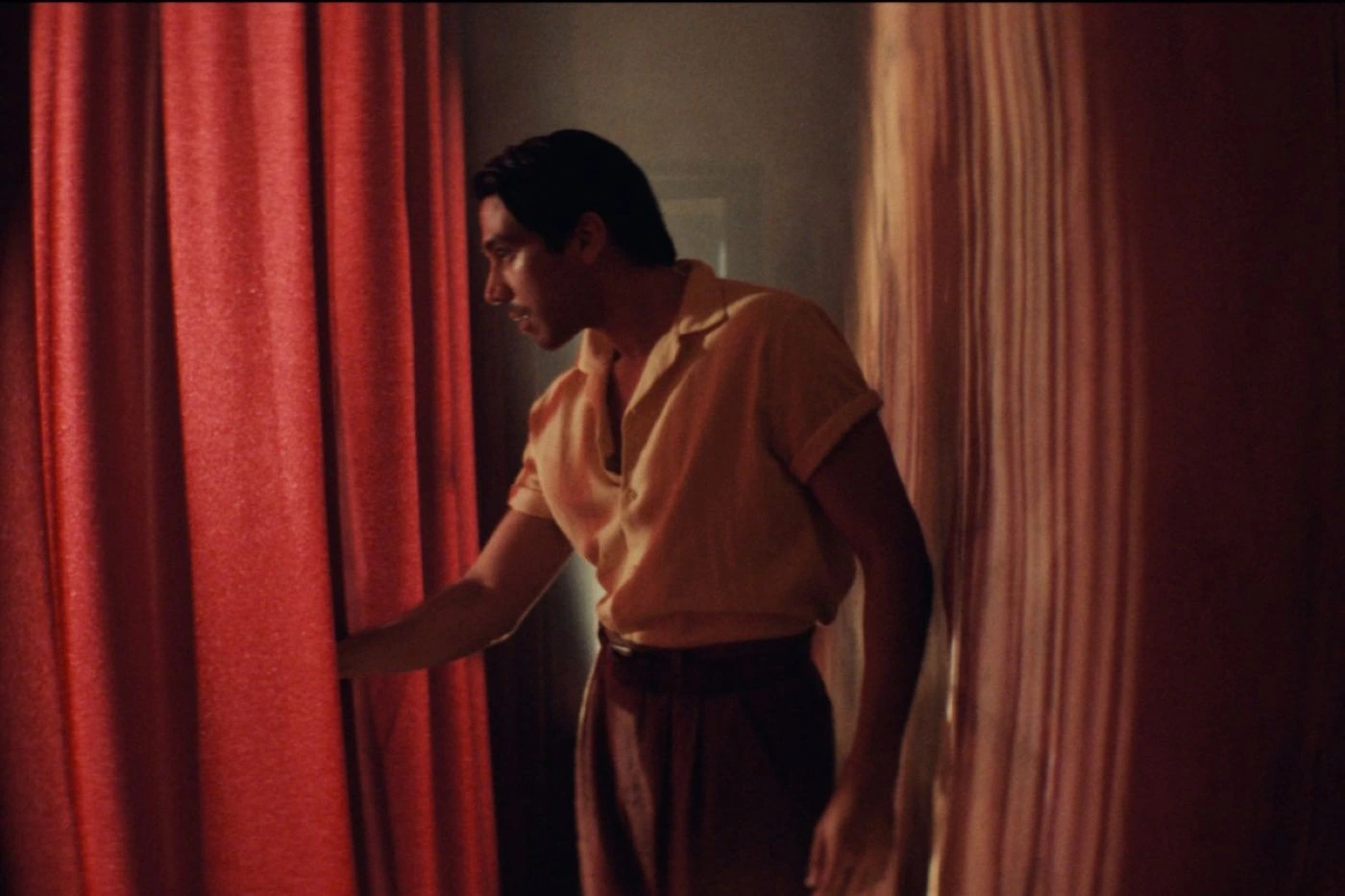 Szenenbild aus dem Film Narciso: Eine Person mit dunklem, zurückgekämmtem Haar trägt ein kurzärmeliges, helles Hemd und eine braune Hose. Sie steht zwischen zwei schweren, roten Vorhängen und hält einen der Vorhänge leicht geöffnet. Das Licht ist warm und gedämpft, wodurch eine intime Atmosphäre entsteht. [erstellt mit KI]