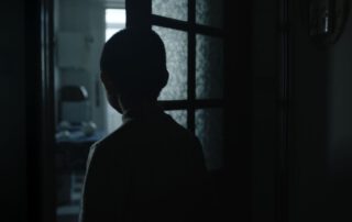 Szenenbild zu Kleptomania: Ein Kind steht mit dem Rücken zur Kamera in einem dunklen Raum und blickt durch eine geöffnete Tür in einen helleren Raum. Die Silhouette des Kindes ist deutlich erkennbar, Details des Gesichts und der Kleidung sind durch die Beleuchtung nicht sichtbar. Im Hintergrund sind ein Schreibtisch und ein Stuhl zu sehen, die von Tageslicht beleuchtet werden. [erstellt mit KI]