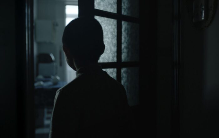 Szenenbild zu Kleptomania: Ein Kind steht mit dem Rücken zur Kamera in einem dunklen Raum und blickt durch eine geöffnete Tür in einen helleren Raum. Die Silhouette des Kindes ist deutlich erkennbar, Details des Gesichts und der Kleidung sind durch die Beleuchtung nicht sichtbar. Im Hintergrund sind ein Schreibtisch und ein Stuhl zu sehen, die von Tageslicht beleuchtet werden. [erstellt mit KI]