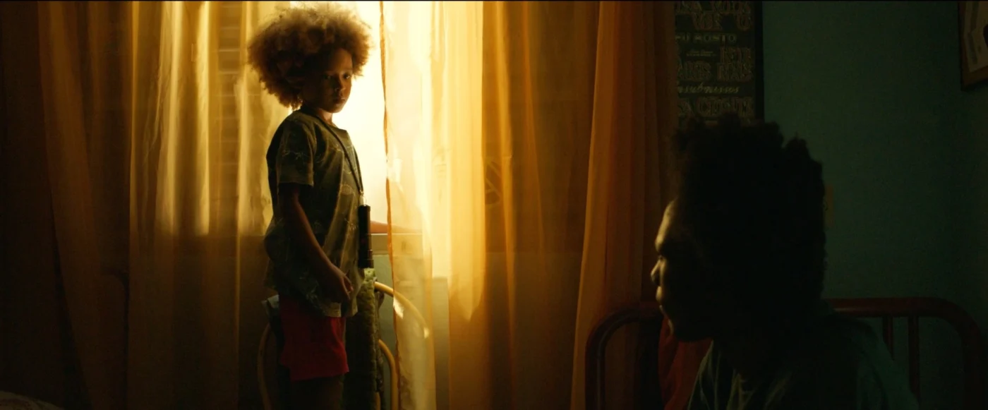 Ein Kind mit lockigem, blondem Haar steht in einem Zimmer vor einem Fenster mit gelbem Vorhang. Das Kind trägt ein graues T-Shirt und rote Shorts. Eine weitere Person sitzt im Halbdunkel auf einem Bett oder Stuhl. Das Licht fällt von hinten durch den Vorhang und taucht die Szene in warmes, goldenes Licht. [erstellt mit KI]