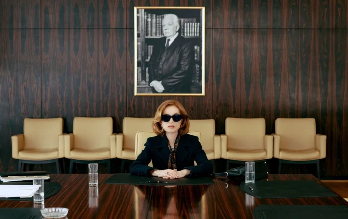 Szenenbild aus "Die reichste Frau der Welt": Eine Frau mit rotbraunen, schulterlangen Haaren sitzt an einem großen Konferenztisch aus dunklem Holz. Sie trägt einen dunklen Blazer und eine Bluse mit Schleife. Vor ihr stehen mehrere Wassergläser und schwarze Schreibunterlagen. Im Hintergrund befinden sich eine Wand mit dunkler Holzvertäfelung, eine Reihe beigefarbener Stühle und ein großes schwarz-weißes Porträt eines älteren Mannes im Anzug, der vor einem Bücherregal steht. [erstellt mit KI]