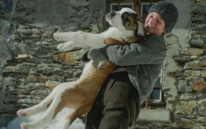 Szenenbild aus Mein Freund Barry: Der junge Georg (Paco von Wyss) in altmodischer Kleidung vor einem Steinhaus mit Barry, einem weiß-braunen Hund im Arm.