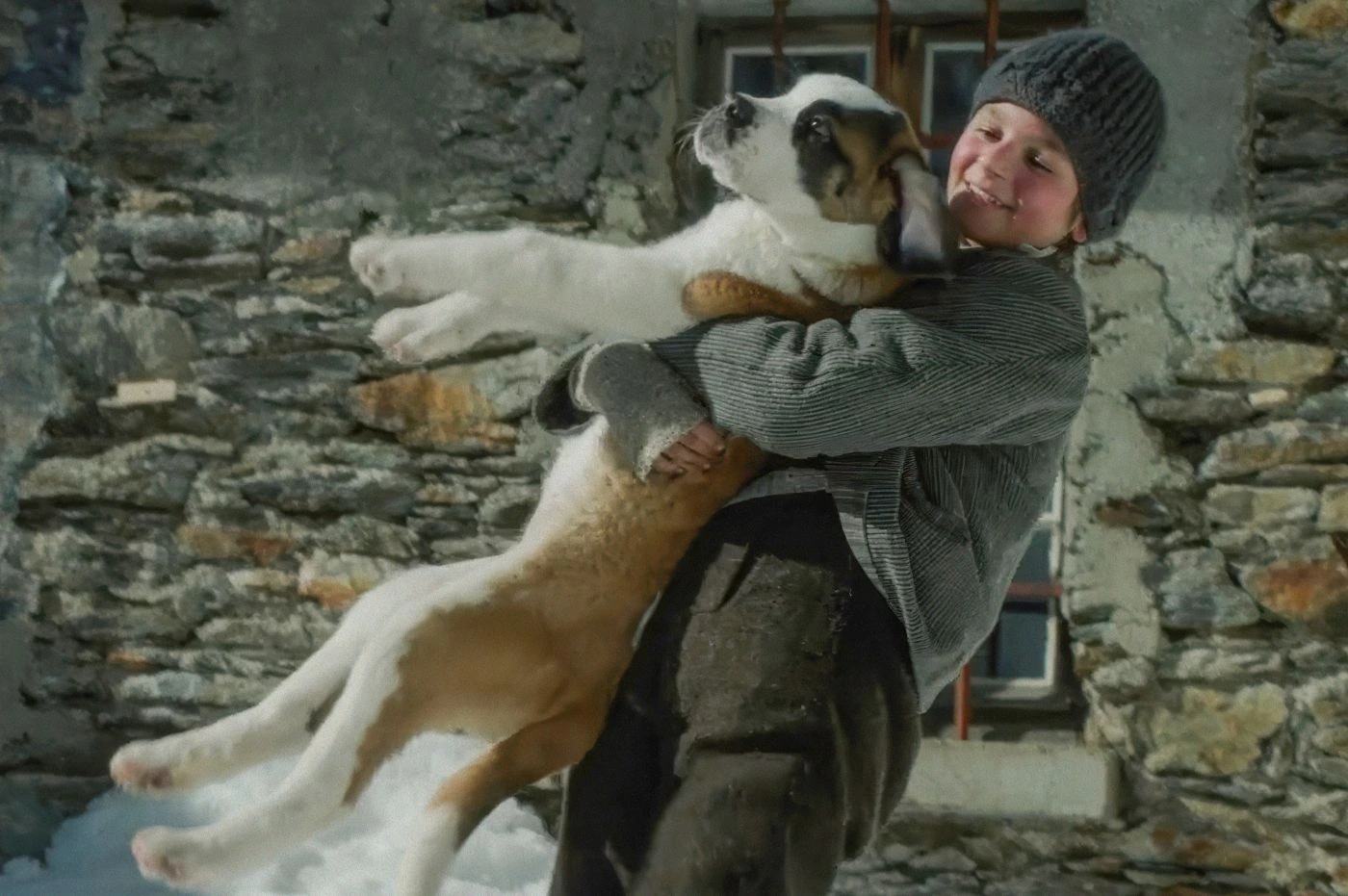 Szenenbild aus Mein Freund Barry: Der junge Georg (Paco von Wyss) in altmodischer Kleidung vor einem Steinhaus mit Barry, einem weiß-braunen Hund im Arm.