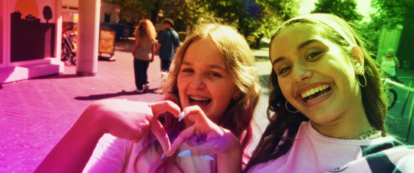 Zwei Personen stehen im Vordergrund und formen mit ihren Händen ein Herz. Beide tragen lässige Kleidung, eine Person trägt große Creolen. Die Szene ist in kräftige, bunte Farben getaucht, wodurch ein auffälliger Farbeffekt entsteht. Im Hintergrund sind weitere Menschen auf einem gepflasterten Platz zu sehen, Bäume spenden Schatten. [erstellt mit KI]