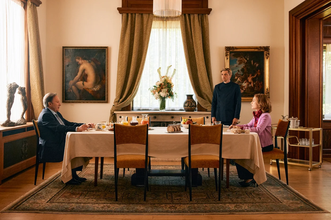 Ein rechteckiger Esstisch steht mittig in einem elegant eingerichteten Raum auf einem großen Teppich. Drei Personen sitzen beziehungsweise stehen an verschiedenen Seiten des Tisches, der mit Geschirr, Gläsern und Speisen gedeckt ist. Die Wände sind mit zwei großen Gemälden und schweren Vorhängen geschmückt. Im Hintergrund steht eine große Blumenvase auf einem Sideboard zwischen den Fenstern. Die Atmosphäre wirkt formell und ruhig. [erstellt mit KI]