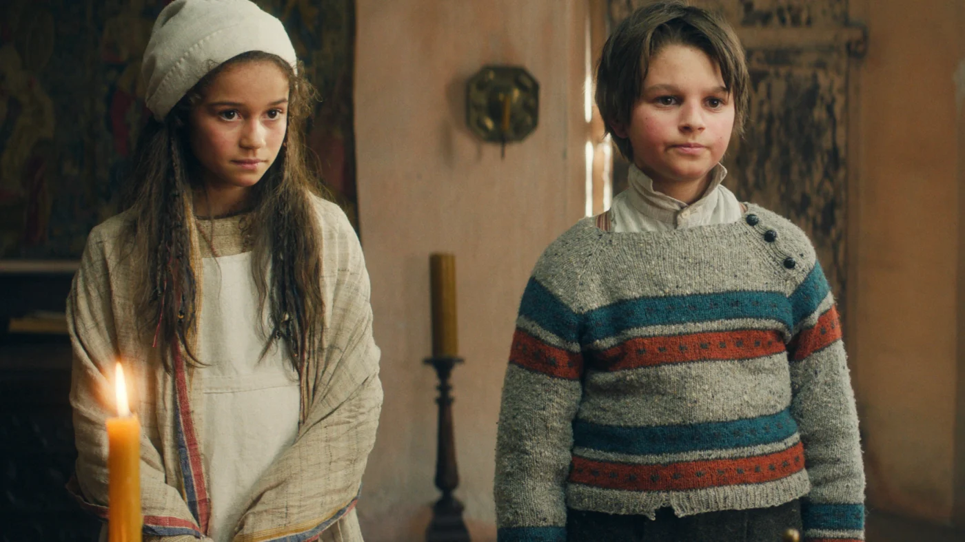 Zwei Kinder stehen nebeneinander in einem Innenraum. Das Kind links trägt eine helle Mütze, ein langes Oberteil und einen Umhang mit Streifenmuster. Das Kind rechts trägt einen grob gestrickten Pullover mit Querstreifen in Blau, Rot und Grau sowie ein weißes Hemd darunter. Im Vordergrund brennt eine Kerze. [erstellt mit KI]