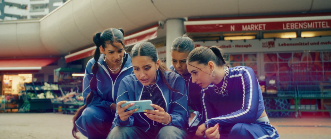 Vier Personen in blauen Trainingsanzügen sitzen eng beieinander auf dem Boden vor einem Supermarkt mit rotem Schriftzug. Sie schauen gemeinsam auf ein hellblaues Smartphone, das eine von ihnen in den Händen hält. Im Hintergrund sind ein geschwungenes Betondach und ein mehrstöckiges Gebäude zu sehen. [erstellt mit KI]