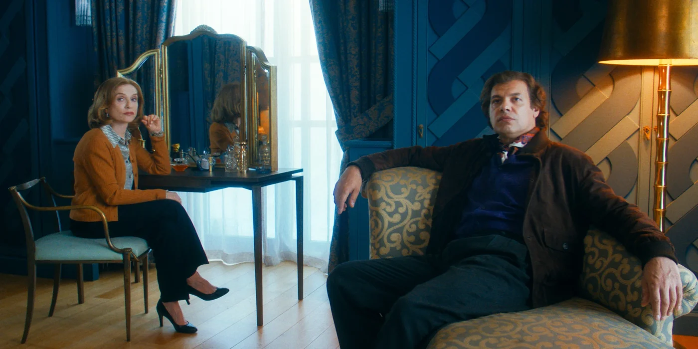 Eine Frau mit blonden, schulterlangen Haaren sitzt an einem Schminktisch mit Spiegel und mehreren Flaschen. Sie trägt einen hellen Blazer, eine dunkle Hose und schwarze Absatzschuhe. Im Vordergrund sitzt ein Mann mit dunklem Sakko und Hemd auf einem gemusterten Sofa. Der Raum ist in Blautönen gehalten, mit schweren Vorhängen, einem Stehlampe mit goldenem Schirm und Parkettboden. Tageslicht fällt durch ein Fenster mit halbtransparentem Vorhang. [erstellt mit KI]