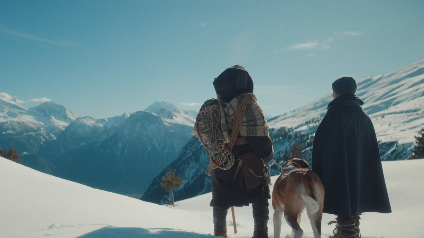 Zwei Personen in dicken Wintermänteln und ein großer Hund stehen auf einem verschneiten Berghang. Die Personen blicken in Richtung eines weiten Tals mit schneebedeckten Bergen im Hintergrund. [erstellt mit KI]