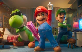 Szenenbild aus DER SUPER MARIO GALAXY FILM: Im Vordergrund stehen drei animierte Figuren in einem Kinderzimmer. Links befindet sich eine grüne Dinosaurierfigur mit weißem Bauch und orangefarbenen Schuhen. In der Mitte steht eine Figur mit roter Mütze und rotem Oberteil, die einen Arm nach oben streckt und eine pink gestreifte Geschenkbox hält. Rechts steht eine Figur mit grüner Mütze und grüner Kleidung, die die Arme verschränkt. Im Hintergrund sind ein Bett mit rot-weiß kariertem Bezug, ein Schreibtisch mit Monitor und verschiedene Möbelstücke zu sehen. [erstellt mit KI]