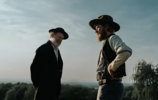 Szenenbild aus A Prayer for the Dying: Zwei Personen stehen im Freien vor einem weiten, leicht bewölkten Himmel. Beide tragen breite Hüte und Kleidung im Stil des 19. Jahrhunderts, darunter Westen, Hemden und lange Mäntel. Die Person links hält die Hände hinter dem Rücken, während die Person rechts eine Hand in die Hüfte stemmt. Im Hintergrund sind Bäume und eine offene Landschaft zu sehen. [erstellt mit KI]