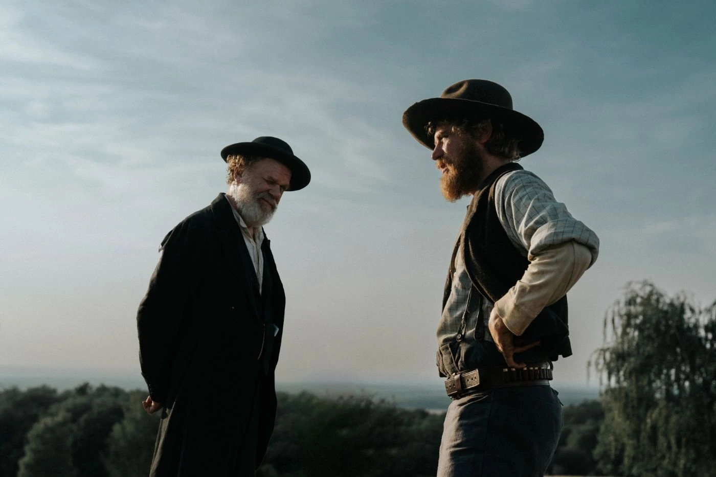Szenenbild aus A Prayer for the Dying: Zwei Personen stehen im Freien vor einem weiten, leicht bewölkten Himmel. Beide tragen breite Hüte und Kleidung im Stil des 19. Jahrhunderts, darunter Westen, Hemden und lange Mäntel. Die Person links hält die Hände hinter dem Rücken, während die Person rechts eine Hand in die Hüfte stemmt. Im Hintergrund sind Bäume und eine offene Landschaft zu sehen. [erstellt mit KI]