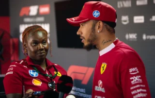 Bild aus Formel 1: Drive to Survive Staffel 8: Zwei Personen stehen nebeneinander vor einer Wand mit zahlreichen Sponsorenlogos. Eine Person trägt ein rotes Ferrari-Teamshirt und eine passende Kappe, auf dem Oberarm sind mehrere Tätowierungen sichtbar. Die andere Person hält ein Mikrofon und trägt ein Oberteil mit verschiedenen Sponsorenaufnähern. Im Hintergrund sind unter anderem die Logos von Ferrari, Shell, AWS und Pirelli zu erkennen. [erstellt mit KI]