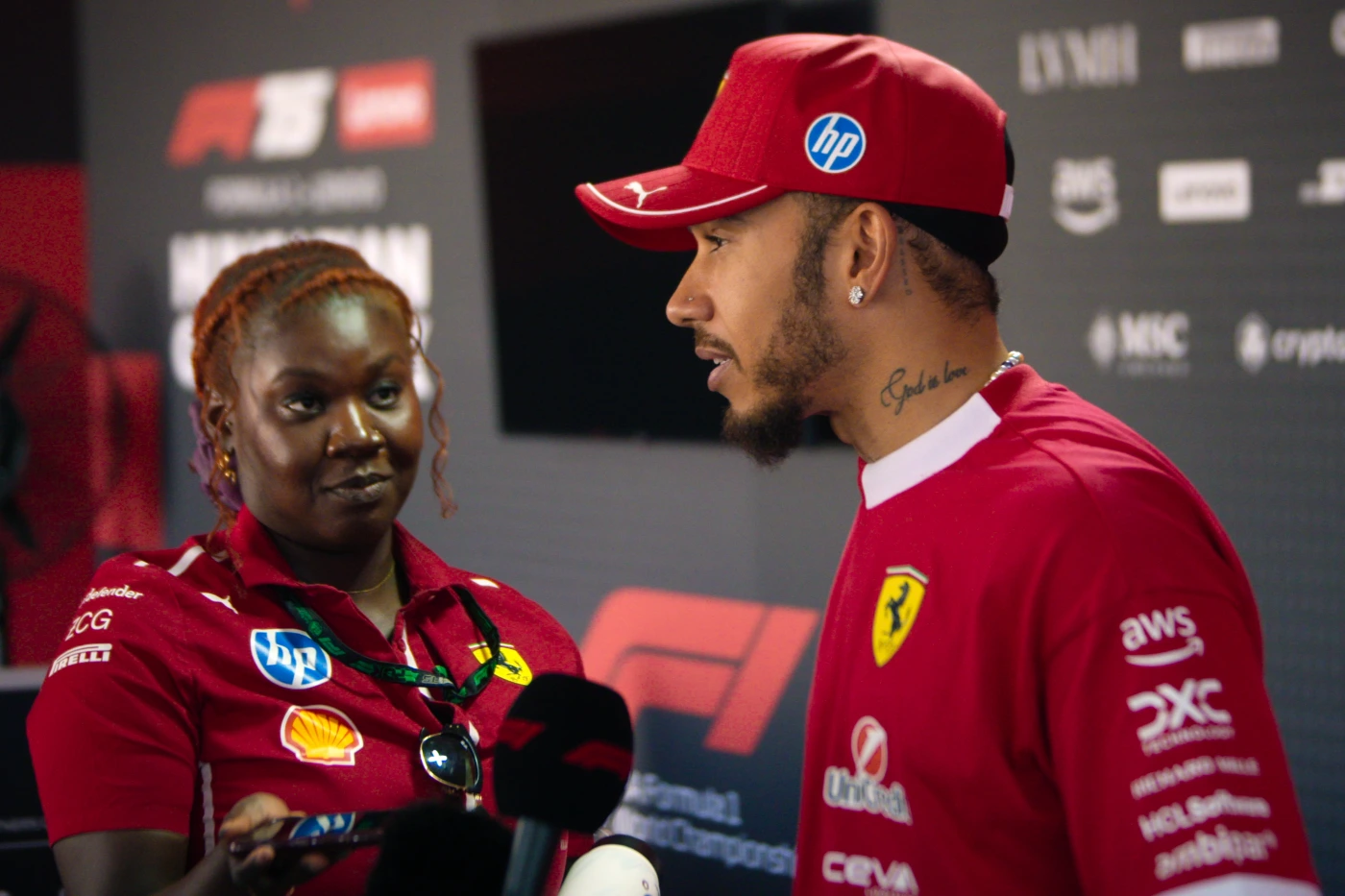 Bild aus Formel 1: Drive to Survive Staffel 8: Zwei Personen stehen nebeneinander vor einer Wand mit zahlreichen Sponsorenlogos. Eine Person trägt ein rotes Ferrari-Teamshirt und eine passende Kappe, auf dem Oberarm sind mehrere Tätowierungen sichtbar. Die andere Person hält ein Mikrofon und trägt ein Oberteil mit verschiedenen Sponsorenaufnähern. Im Hintergrund sind unter anderem die Logos von Ferrari, Shell, AWS und Pirelli zu erkennen. [erstellt mit KI]