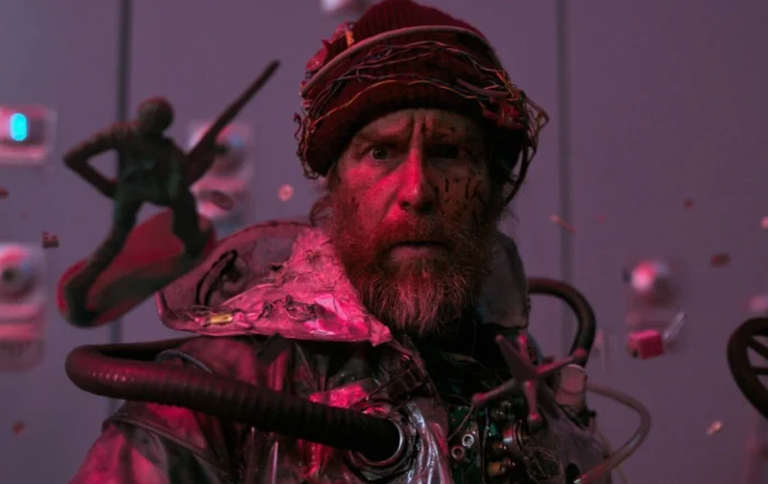 Szenenbild aus Good Luck, Have Fun, Don't Die: Eine Person trägt einen roten Wollhut und eine metallisch glänzende Jacke mit zahlreichen technischen Komponenten, Kabeln und Schläuchen. Im Vordergrund schwebt eine kleine, grüne Figur mit Schwert und Schild. Der Hintergrund zeigt eine Wand mit mehreren runden Bedienelementen und einer violetten Beleuchtung. [erstellt mit KI]