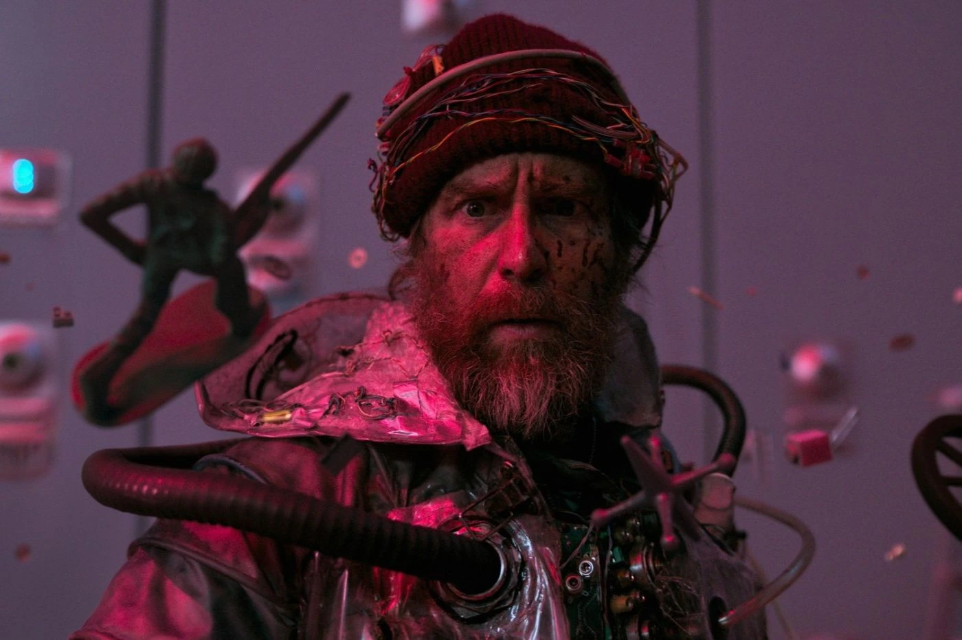 Szenenbild aus Good Luck, Have Fun, Don't Die: Eine Person trägt einen roten Wollhut und eine metallisch glänzende Jacke mit zahlreichen technischen Komponenten, Kabeln und Schläuchen. Im Vordergrund schwebt eine kleine, grüne Figur mit Schwert und Schild. Der Hintergrund zeigt eine Wand mit mehreren runden Bedienelementen und einer violetten Beleuchtung. [erstellt mit KI]