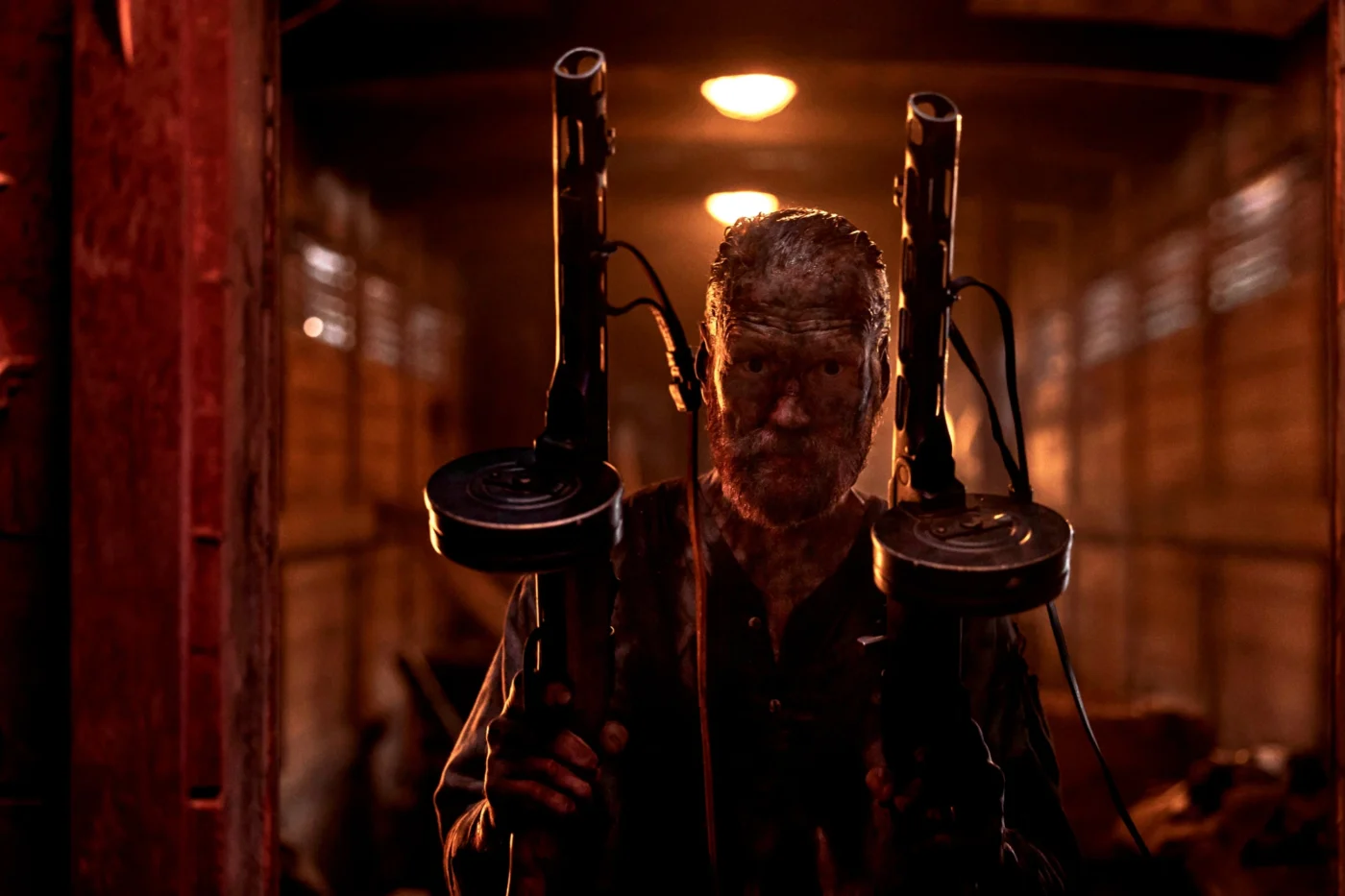 Eine Person hält zwei große, trommelmagazinartige Waffen mit langen Kabeln, die nach oben führen. Die Szene spielt in einem schwach beleuchteten, hölzernen Waggon mit warmem, rötlichem Licht. Im Hintergrund sind diffuse Lichtquellen an der Decke sichtbar. [erstellt mit KI]