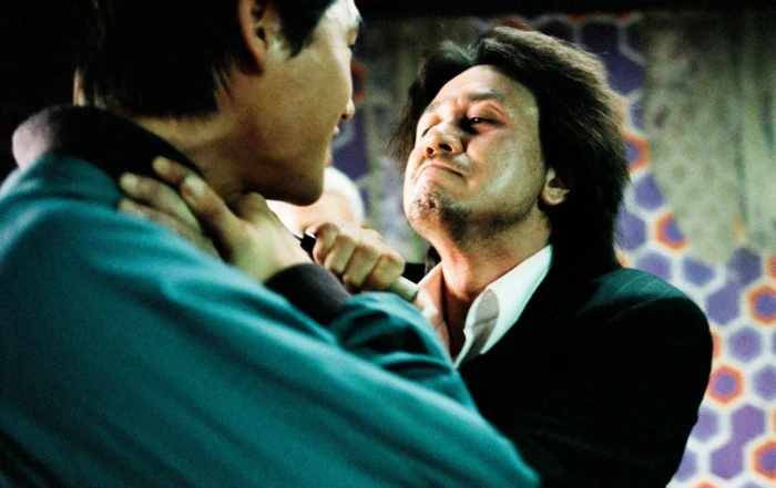 Szenenbild aus Oldboy: Zwei Männer stehen sich in einem Innenraum gegenüber. Einer trägt ein dunkles Jackett und ein weißes Hemd, der andere ein türkisfarbenes Oberteil. Der Mann im Jackett wird am Kragen gepackt. Im Hintergrund ist eine Wand mit geometrischem Muster in Lila-, Orange- und Grautönen zu sehen. [erstellt mit KI]