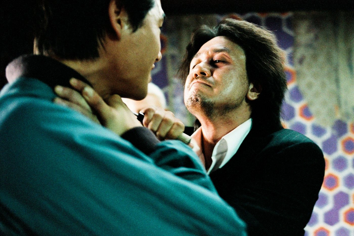 Szenenbild aus Oldboy: Zwei Männer stehen sich in einem Innenraum gegenüber. Einer trägt ein dunkles Jackett und ein weißes Hemd, der andere ein türkisfarbenes Oberteil. Der Mann im Jackett wird am Kragen gepackt. Im Hintergrund ist eine Wand mit geometrischem Muster in Lila-, Orange- und Grautönen zu sehen. [erstellt mit KI]