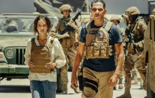 Szenenbild aus Operation: Kabul: Im Vordergrund stehen zwei Personen mit sandfarbenen Schutzwesten. Die Person links trägt ein weißes Hemd, die Person rechts ein dunkelblaues T-Shirt mit einem runden Aufnäher auf der Brust. Im Hintergrund sind mehrere Soldaten in Tarnuniformen und Helmen zu sehen, die Gewehre tragen. Ein Militärfahrzeug mit offenem Dach steht hinter den Personen. Die Szene spielt bei Tageslicht, die Umgebung wirkt staubig und trocken. [erstellt mit KI]