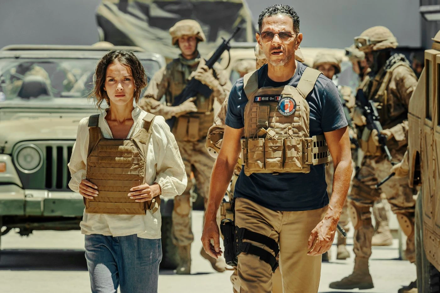 Szenenbild aus Operation: Kabul: Im Vordergrund stehen zwei Personen mit sandfarbenen Schutzwesten. Die Person links trägt ein weißes Hemd, die Person rechts ein dunkelblaues T-Shirt mit einem runden Aufnäher auf der Brust. Im Hintergrund sind mehrere Soldaten in Tarnuniformen und Helmen zu sehen, die Gewehre tragen. Ein Militärfahrzeug mit offenem Dach steht hinter den Personen. Die Szene spielt bei Tageslicht, die Umgebung wirkt staubig und trocken. [erstellt mit KI]