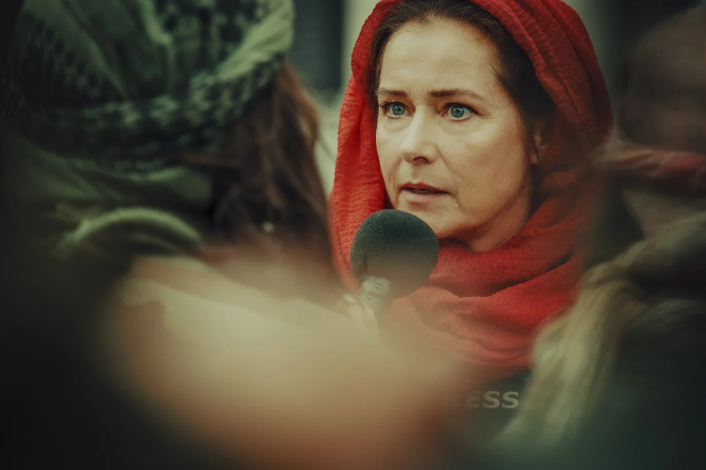 Eine Person mit rotem Kopftuch steht im Vordergrund und spricht in ein Mikrofon. Die Szene wirkt unscharf und ist teilweise durch einen verschwommenen Vordergrund verdeckt. [erstellt mit KI]