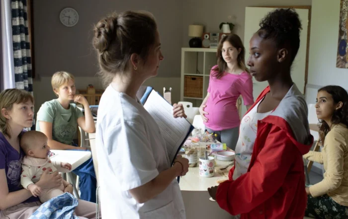 Szenenbild aus Jeunes meres - Junge Mütter: In einem hellen Raum mit schlichter Einrichtung stehen mehrere Frauen und Kinder zusammen. Im Vordergrund hält eine Person in weißer Kleidung ein Klemmbrett und spricht mit einer anderen Person im roten Oberteil. Im Hintergrund sitzt eine Frau mit einem Baby auf dem Schoß neben einer weiteren erwachsenen Person. Auf dem Tisch in der Mitte stehen verschiedene Gegenstände, darunter Becher und Verpackungen. [erstellt mit KI]