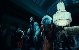 Szenenbild aus The Bride!: Eine Gruppe von Menschen tanzt in einem prunkvollen Saal mit hoher Decke und großem Kronleuchter. Im Hintergrund stehen Blumenarrangements und ein Vorhang mit Falten. Die Szene ist dunkel gehalten, das Licht des Kronleuchters dominiert den Raum. Die Kleidung der Personen ist auffällig und elegant, darunter ein rotes Kleid und ein gestreifter Anzug.