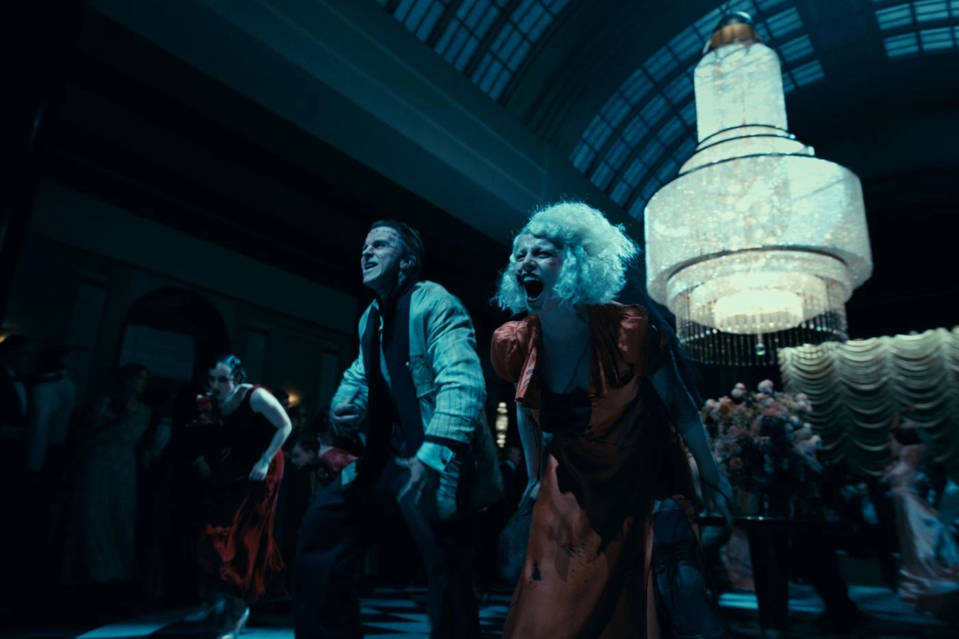 Szenenbild aus The Bride!: Eine Gruppe von Menschen tanzt in einem prunkvollen Saal mit hoher Decke und großem Kronleuchter. Im Hintergrund stehen Blumenarrangements und ein Vorhang mit Falten. Die Szene ist dunkel gehalten, das Licht des Kronleuchters dominiert den Raum. Die Kleidung der Personen ist auffällig und elegant, darunter ein rotes Kleid und ein gestreifter Anzug.