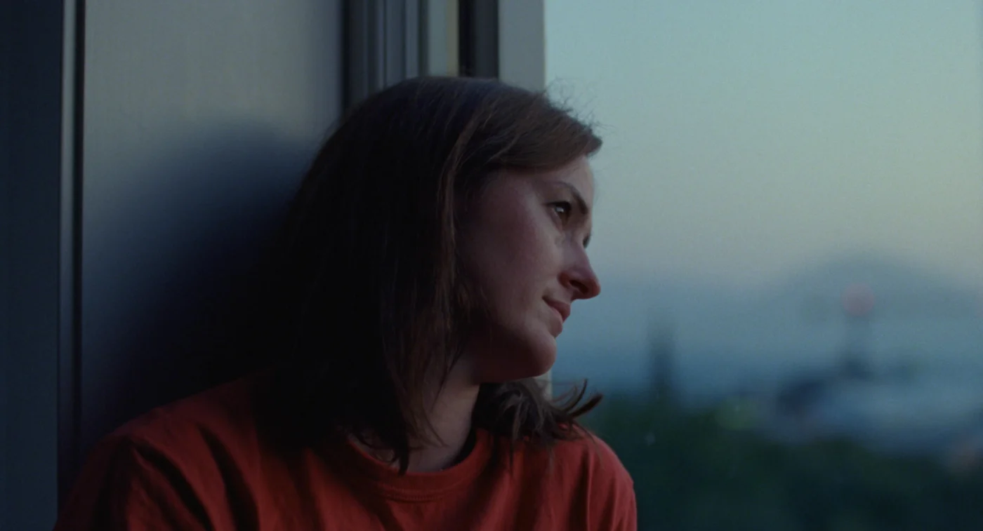 Eine Frau im roten Pullover lehnt mit dem Kopf gegen eine Fensterscheibe.