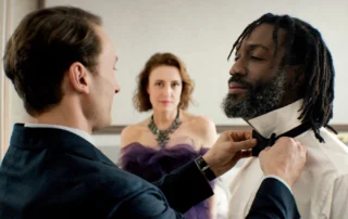 Szenenbild aus "Gavagai": Ein Mann im Anzug bindet einem anderen Mann mit Dreadlocks eine schwarze Fliege. Im Hintergrund steht eine Frau in einem schulterfreien, lilafarbenen Kleid mit auffälliger Halskette. Die Szene spielt in einem hellen Raum mit neutraler Wandfarbe.