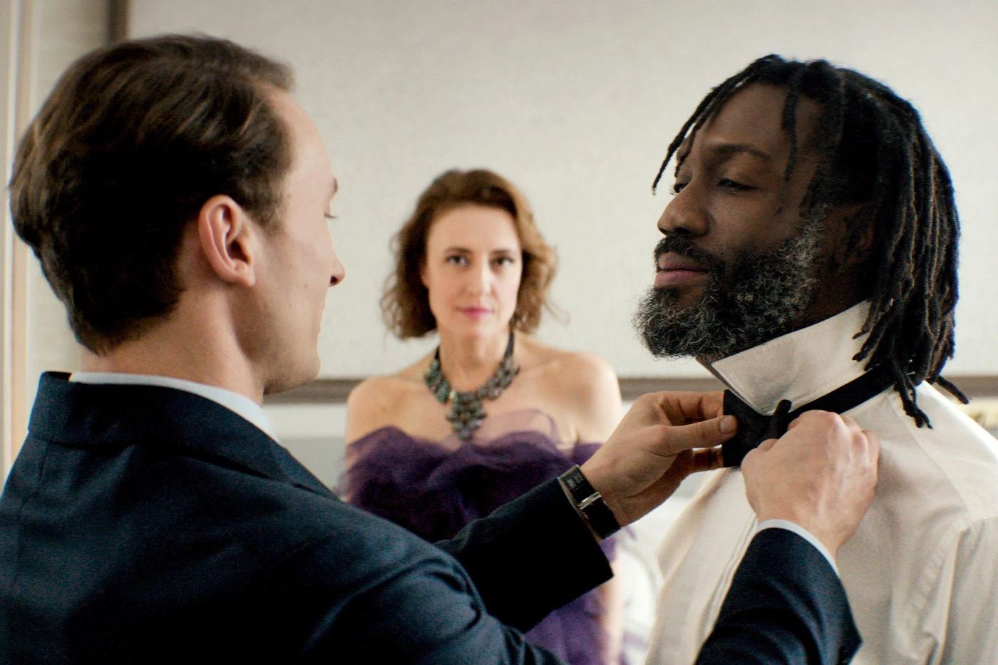 Szenenbild aus "Gavagai": Ein Mann im Anzug bindet einem anderen Mann mit Dreadlocks eine schwarze Fliege. Im Hintergrund steht eine Frau in einem schulterfreien, lilafarbenen Kleid mit auffälliger Halskette. Die Szene spielt in einem hellen Raum mit neutraler Wandfarbe.
