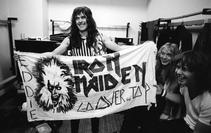Szenenbild aus Iron Maiden - Burning Ambition: Eine Person mit langen Haaren hält ein großes, handbemaltes Banner in einem Raum mit sichtbaren Kleiderständern und Kisten. Das Banner zeigt links ein gezeichnetes Gesicht mit wilden Haaren und weit aufgerissenen Augen. Rechts daneben stehen in großen, markanten Buchstaben mehrere Wörter. Zwei weitere Personen sitzen rechts am Bildrand, eine davon hält das Banner mit fest. Die Szene ist in schwarz-weiß gehalten. [erstellt mit KI]
