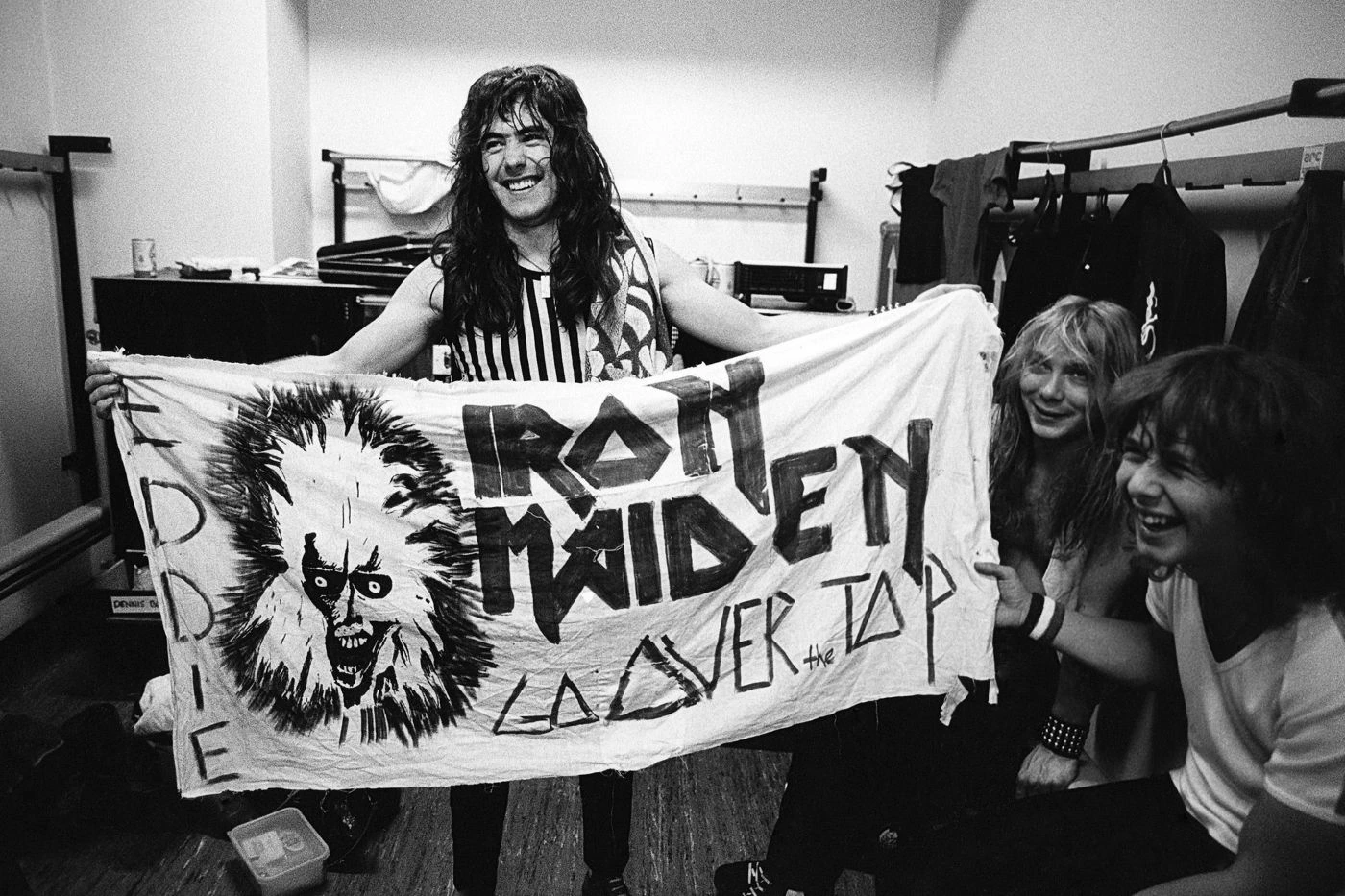 Szenenbild aus Iron Maiden - Burning Ambition: Eine Person mit langen Haaren hält ein großes, handbemaltes Banner in einem Raum mit sichtbaren Kleiderständern und Kisten. Das Banner zeigt links ein gezeichnetes Gesicht mit wilden Haaren und weit aufgerissenen Augen. Rechts daneben stehen in großen, markanten Buchstaben mehrere Wörter. Zwei weitere Personen sitzen rechts am Bildrand, eine davon hält das Banner mit fest. Die Szene ist in schwarz-weiß gehalten. [erstellt mit KI]