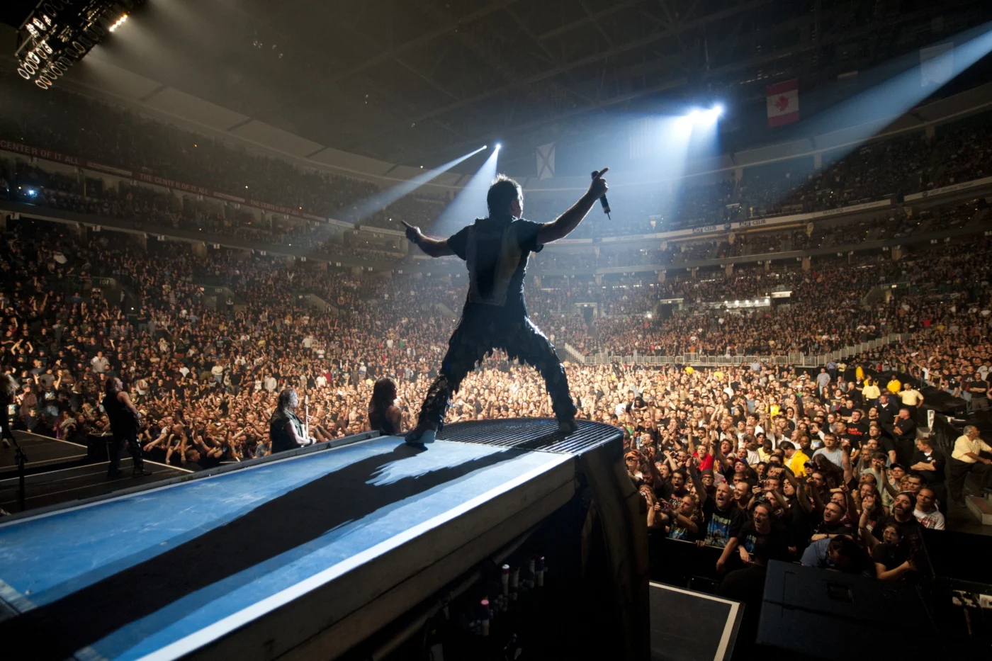 Szenenbild aus Iron Maiden - Burning Ambition: Eine Person steht mit ausgebreiteten Armen auf einer erhöhten Bühnenplattform und blickt in eine große, dicht gefüllte Konzerthalle. Die Person ist von hinten zu sehen und wird von starkem Bühnenlicht angestrahlt, das einen langen Schatten auf die Bühne wirft. Im Hintergrund sind zahlreiche Konzertbesucher zu erkennen, die in Richtung Bühne schauen. Die Halle ist groß und von mehreren Rängen umgeben. [erstellt mit KI]