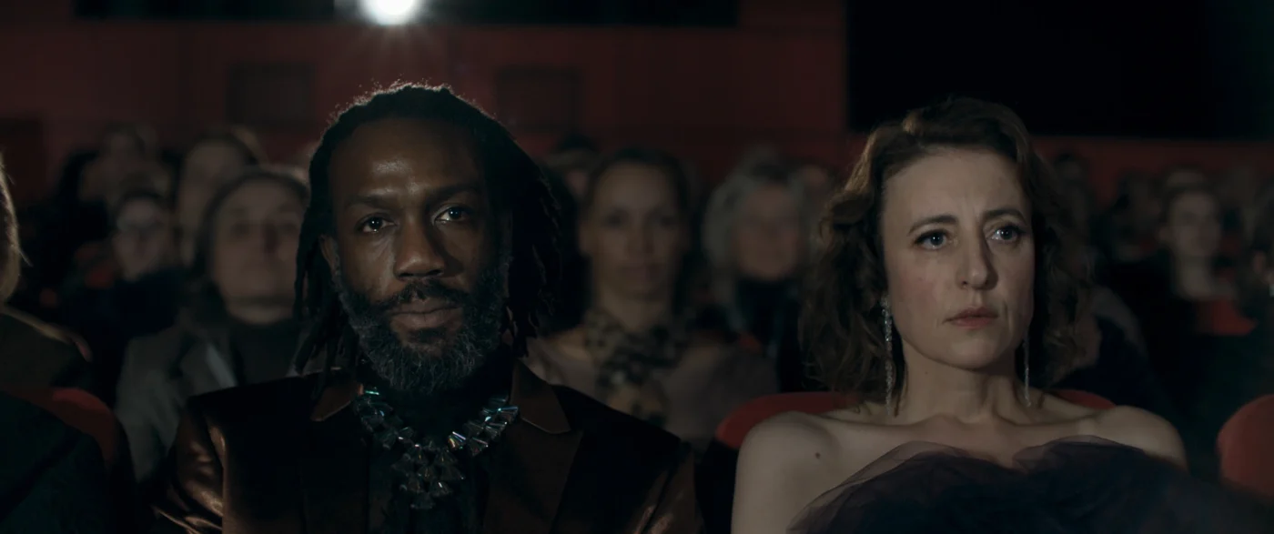 Zwei Personen sitzen in einem Kinosaal in der ersten Reihe. Die linke Person trägt ein auffälliges, glänzendes Oberteil und eine große Halskette. Die rechte Person trägt ein schulterfreies Kleid. Im Hintergrund sind weitere Menschen auf roten Kinosesseln zu sehen. Die Beleuchtung ist gedämpft, im hinteren Bereich ist eine Lichtquelle sichtbar. [erstellt mit KI]