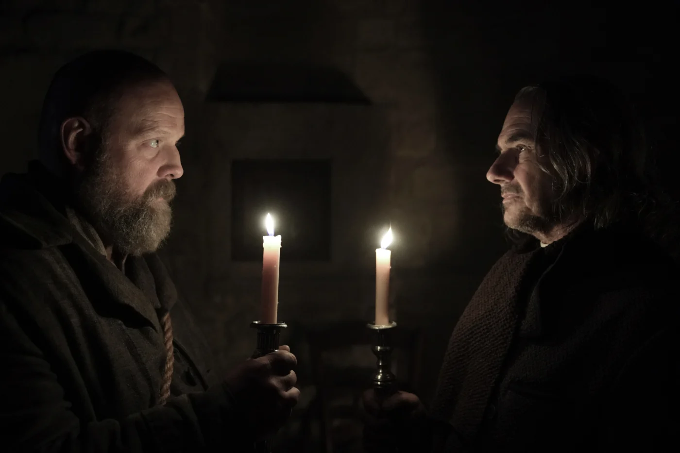 Zwei Männer stehen sich in einem Kellergewölbe mit Kerzen in den Händen gegenüber. Die Szenerie wird nur vom Kerzenlicht erhellt.