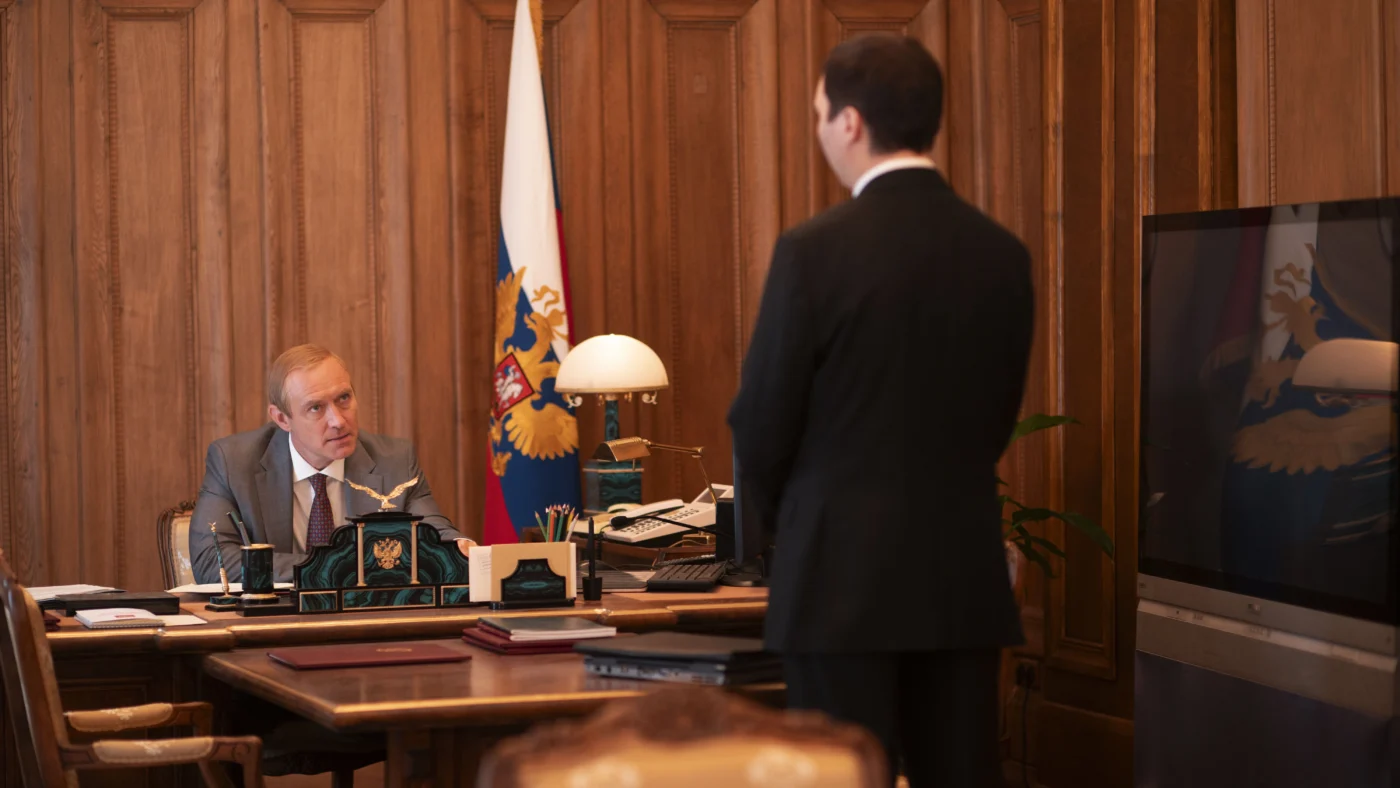 Ein Mann im Anzug steht mit dem Rücken zur Kamera vor einem großen Schreibtisch hinter dem ein weiterer Mann sitzt. Links neben dem sitzenden Mann steht eine russische Flagge.