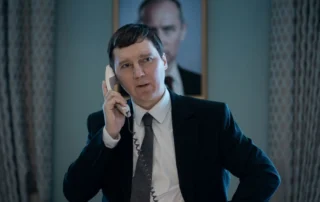 Szenenbild aus Der Magier im Kreml: Ein Mann im Anzug und Schlips am Telefon vor einem Porträt Putins.