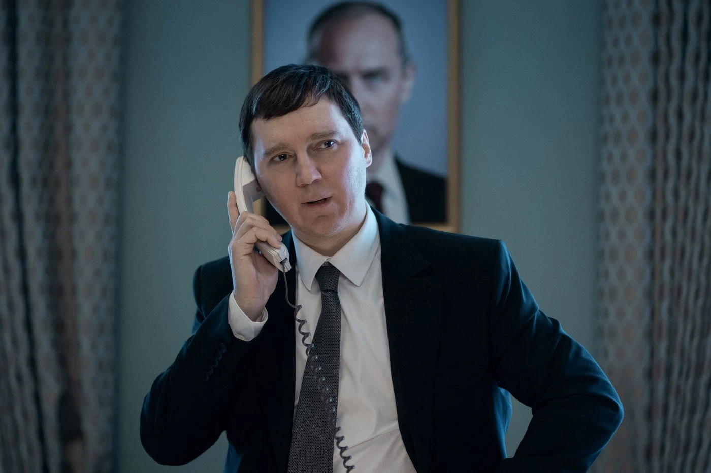 Szenenbild aus Der Magier im Kreml: Ein Mann im Anzug und Schlips am Telefon vor einem Porträt Putins.