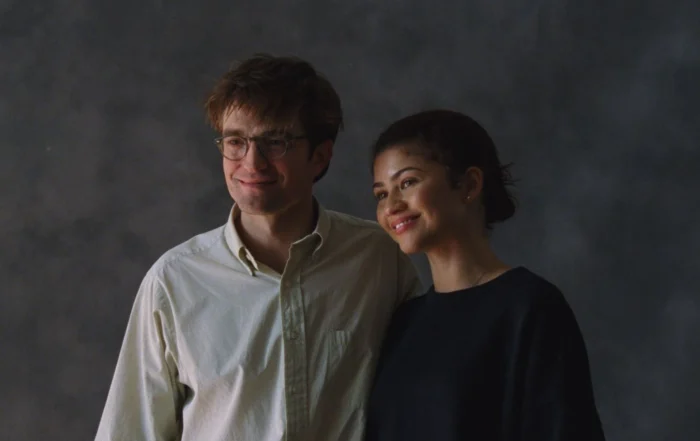Szenenbild aus Das Drama: Ein junger Mann und eine junge Frau stehen vor einem dunkelgrauen Hintergrund und lächeln. Er trägt ein weißes Hemd, sie ein schwarzes Oberteil.