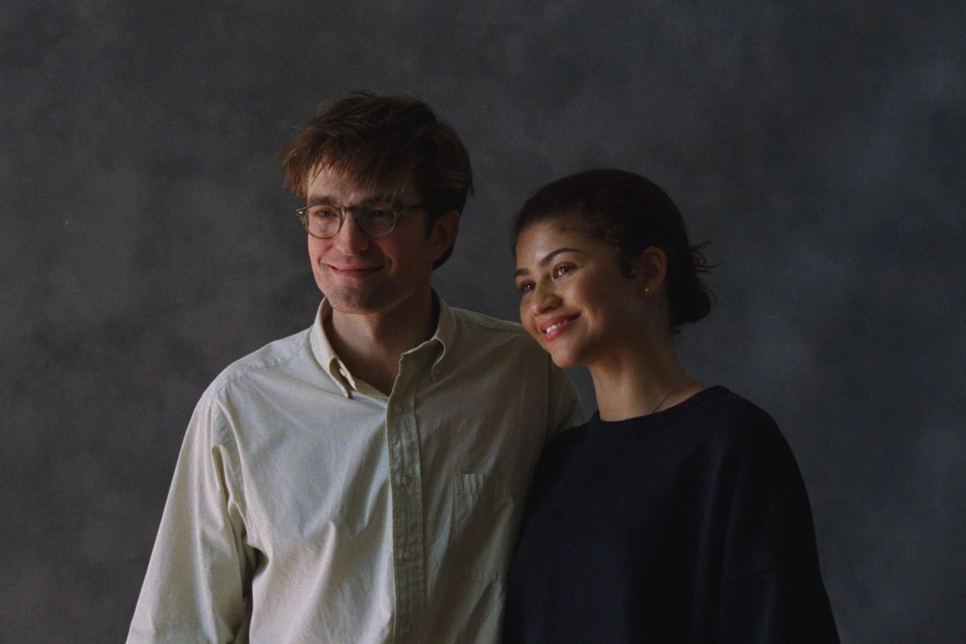Szenenbild aus Das Drama: Ein junger Mann und eine junge Frau stehen vor einem dunkelgrauen Hintergrund und lächeln. Er trägt ein weißes Hemd, sie ein schwarzes Oberteil.