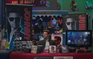 Szenenbild aus Mortal Kombat II: Ein Mann sitzt an einem Tisch, der mit verschiedenen Fanartikeln und DVDs bedeckt ist. Hinter ihm stehen zwei große, senkrechte Banner mit dem Namen 'JOHNNY' und dem Porträt eines Mannes. Rechts auf dem Tisch steht ein Flachbildschirm, auf dem eine Kampfszene aus einem Film oder Videospiel zu sehen ist. Links auf dem Tisch liegen mehrere glänzende Gegenstände, die wie Requisiten oder Nachbildungen von Waffen aussehen. [erstellt mit KI]