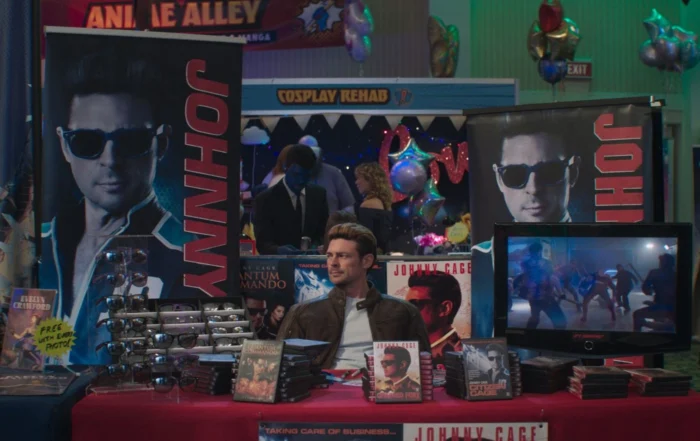 Szenenbild aus Mortal Kombat II: Ein Mann sitzt an einem Tisch, der mit verschiedenen Fanartikeln und DVDs bedeckt ist. Hinter ihm stehen zwei große, senkrechte Banner mit dem Namen 'JOHNNY' und dem Porträt eines Mannes. Rechts auf dem Tisch steht ein Flachbildschirm, auf dem eine Kampfszene aus einem Film oder Videospiel zu sehen ist. Links auf dem Tisch liegen mehrere glänzende Gegenstände, die wie Requisiten oder Nachbildungen von Waffen aussehen. [erstellt mit KI]