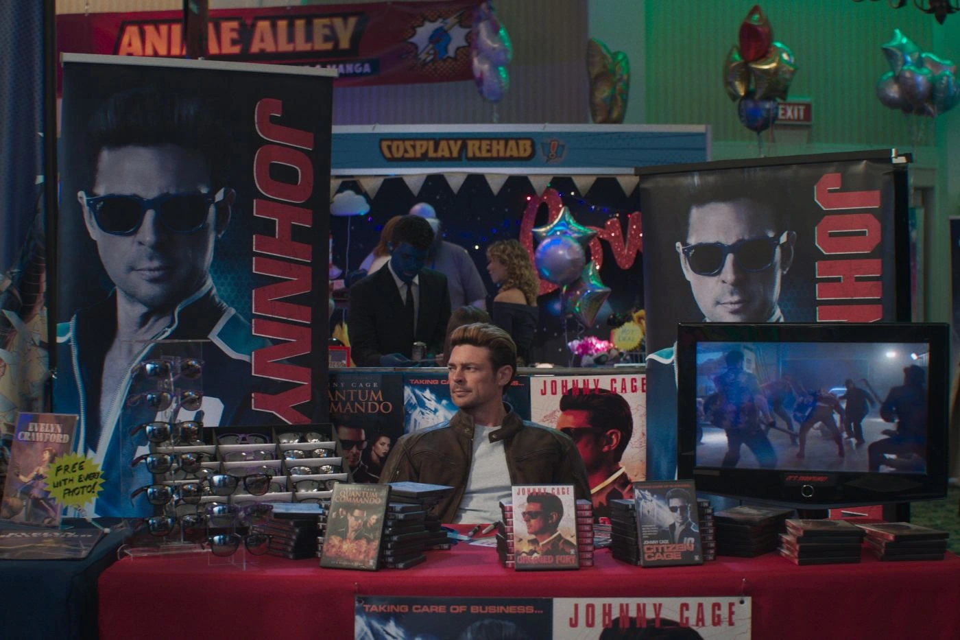 Szenenbild aus Mortal Kombat II: Ein Mann sitzt an einem Tisch, der mit verschiedenen Fanartikeln und DVDs bedeckt ist. Hinter ihm stehen zwei große, senkrechte Banner mit dem Namen 'JOHNNY' und dem Porträt eines Mannes. Rechts auf dem Tisch steht ein Flachbildschirm, auf dem eine Kampfszene aus einem Film oder Videospiel zu sehen ist. Links auf dem Tisch liegen mehrere glänzende Gegenstände, die wie Requisiten oder Nachbildungen von Waffen aussehen. [erstellt mit KI]