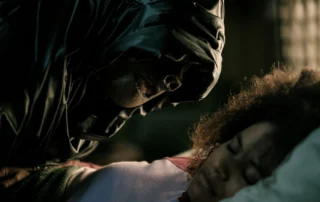 Eine Person mit dunkler Kapuze beugt sich dicht über eine andere Person, deren Gesicht nicht sichtbar ist. Die Szene ist düster ausgeleuchtet, der Fokus liegt auf dem bedrohlichen Gesichtsausdruck der Person mit Kapuze. Das Bild stammt aus dem Film »They Will Kill You«. Im Vordergrund ist ein unscharfer Bereich zu sehen, der Details verdeckt. [erstellt mit KI]