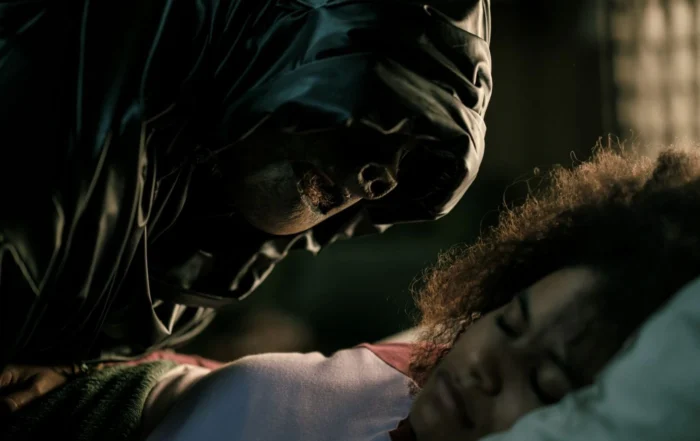 Eine Person mit dunkler Kapuze beugt sich dicht über eine andere Person, deren Gesicht nicht sichtbar ist. Die Szene ist düster ausgeleuchtet, der Fokus liegt auf dem bedrohlichen Gesichtsausdruck der Person mit Kapuze. Das Bild stammt aus dem Film »They Will Kill You«. Im Vordergrund ist ein unscharfer Bereich zu sehen, der Details verdeckt. [erstellt mit KI]