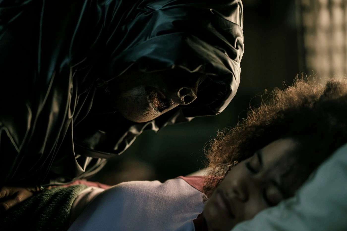 Eine Person mit dunkler Kapuze beugt sich dicht über eine andere Person, deren Gesicht nicht sichtbar ist. Die Szene ist düster ausgeleuchtet, der Fokus liegt auf dem bedrohlichen Gesichtsausdruck der Person mit Kapuze. Das Bild stammt aus dem Film »They Will Kill You«. Im Vordergrund ist ein unscharfer Bereich zu sehen, der Details verdeckt. [erstellt mit KI]