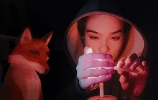 Bild aus A Fox under a Pink Moon: Im Dunkeln zündet eine Frau mit Kapuze eine Kerze an. Ein animierter Fuchs sitzt neben ihr.