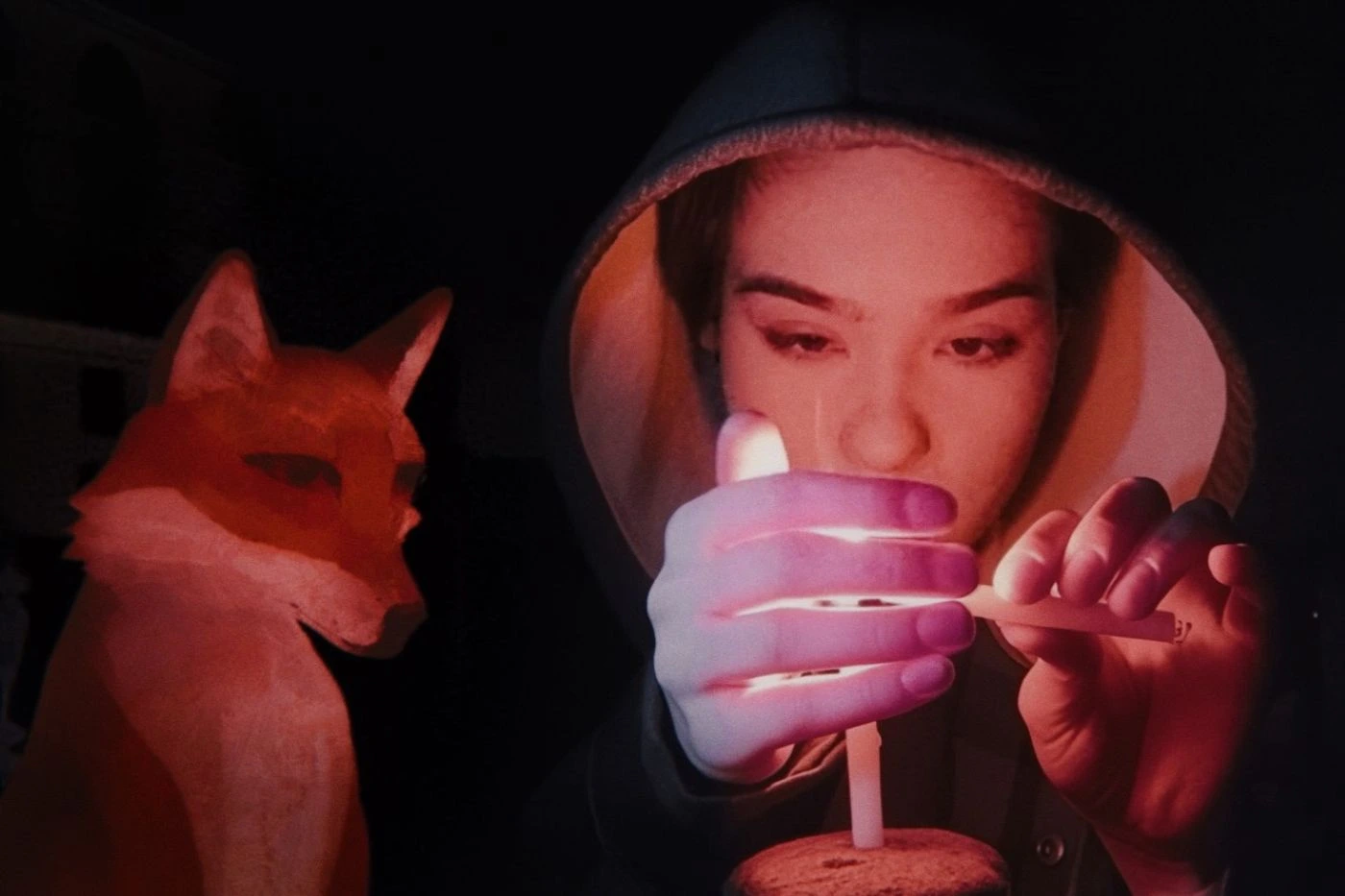 Bild aus A Fox under a Pink Moon: Im Dunkeln zündet eine Frau mit Kapuze eine Kerze an. Ein animierter Fuchs sitzt neben ihr.